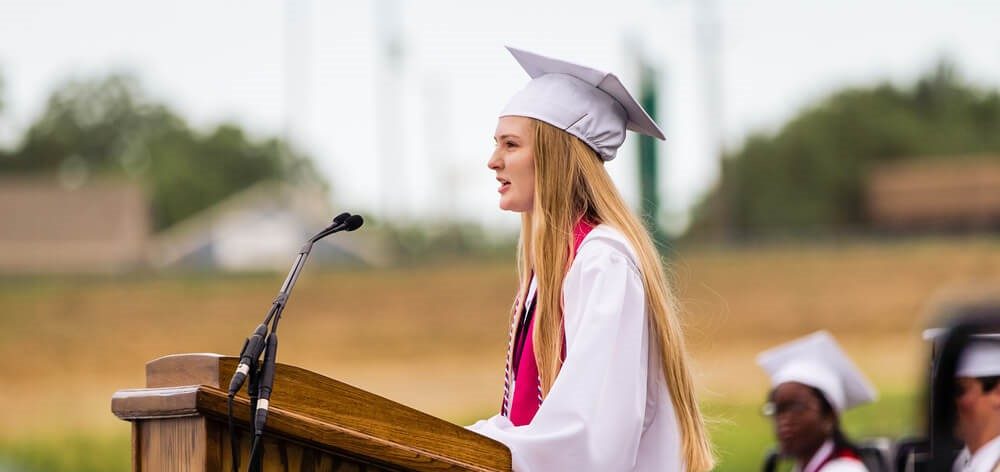 First generation college student wears a white graduation gown while she gives a speech during an outdoor graduation ceremony