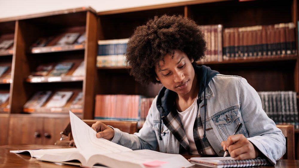 Black male student sitting at a desk searching for scholarships for black students in a book while taking notes in a library
