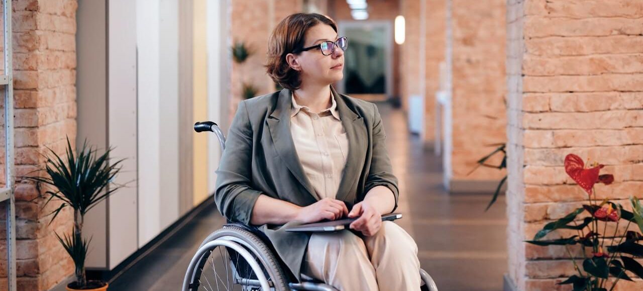 Disabled female parent in wheelchair holding a computer as she sits in an office hallway as she gets ready to go in her office to look for scholarships for students with disabled parent(s).