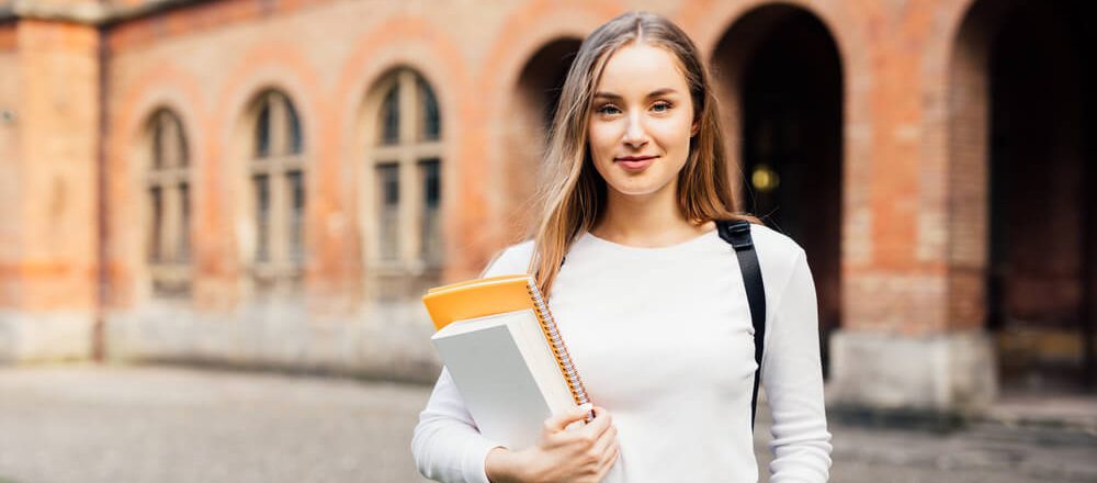 Female student who knows how to decline a college acceptance smiles at the camera and holds her books